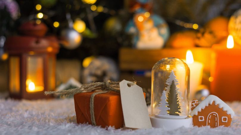Orange gift, with tag, beside a snow globe and small toy house with candles and lanterns in the background.