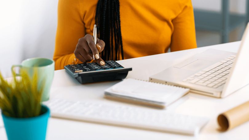 A woman sitting at a desk determining the replacement value of a home using a calculator, laptop and documents.