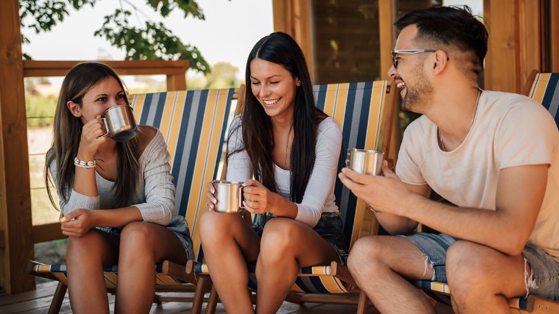 Un homme et deux femmes dans la vingtaine prennent plaisir à déguster un café sous le porche d’un chalet.