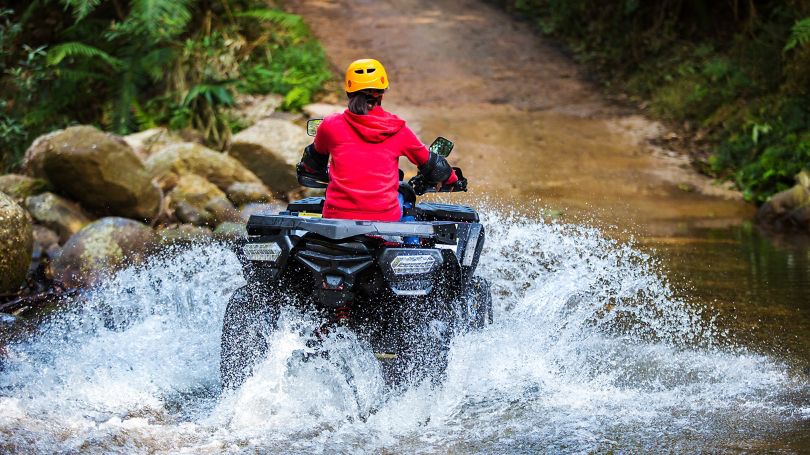A girl in a red hoodie driving an ATV through a stream, creating splashes of water.
