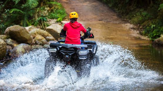 A girl in a red hoodie driving an ATV through a stream, creating splashes of water.