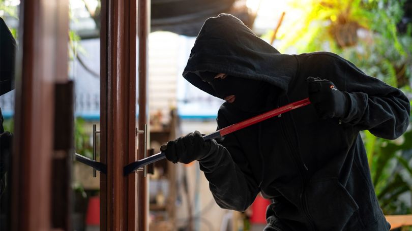 Un individu entièrement vêtu de noir, portant une cagoule et un masque, s’introduit dans une maison.