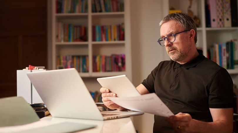 Male sitting at table with laptop looking reviewing paperwork