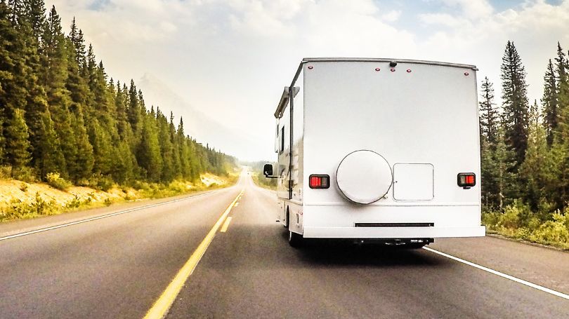 A Class C RV travels down a wilderness highway with fir trees lining the roadway