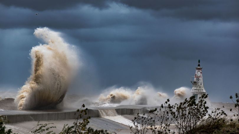 A stormy coastline with waves crashing the rocks in front of a lighthouse
