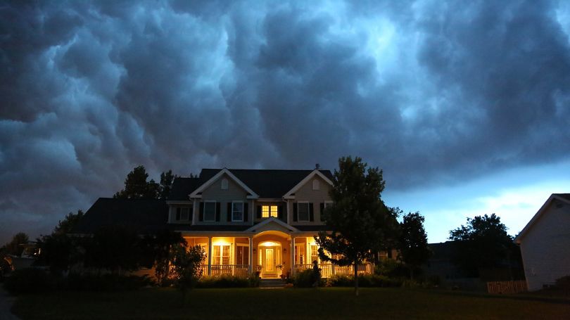 Une maison chaleureusement éclairée se dresse sous un ciel sombre et orageux, entourée d’arbres et de maisons voisines.
