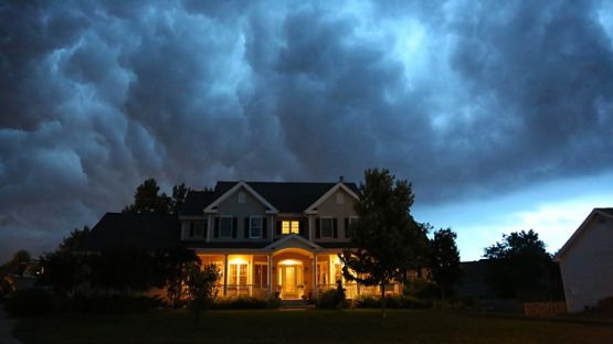 A warmly lit house stands beneath a dark, stormy sky, surrounded by trees and neighboring homes.