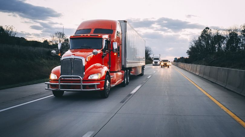 Un semi-remorque roule sur une autoroute au crépuscule, suivi par un autre camion et plusieurs véhicules.