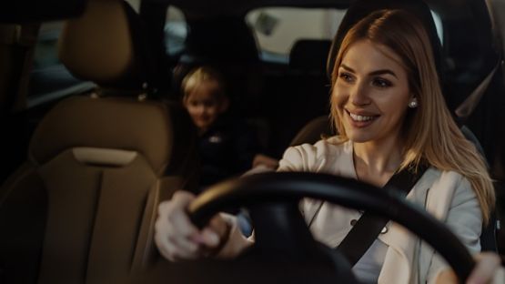 Une femme conduisant une voiture avec un enfant sur la banquette arrière; visages floutés pour respecter la vie privée.