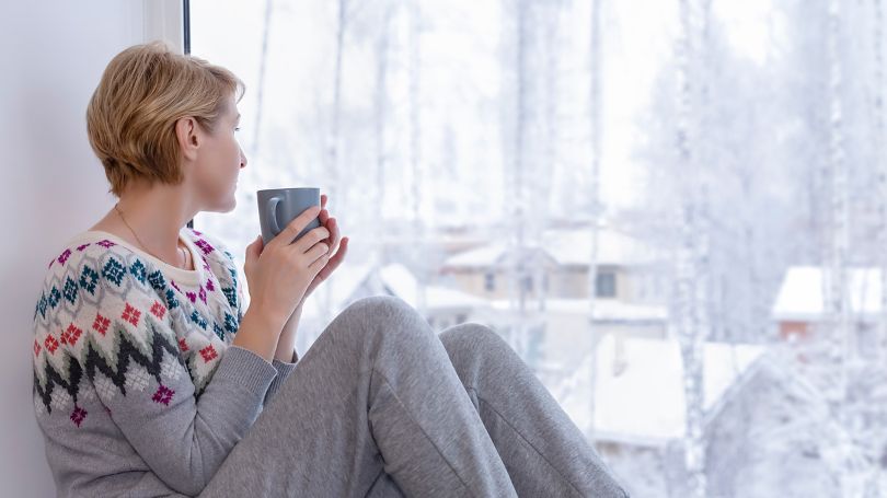 A woman sitting on the windowsill with a cup of tea looking out at her snow-covered neighbourhood.
