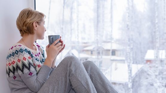 A woman sitting on the windowsill with a cup of tea looking out at her snow-covered neighbourhood.
