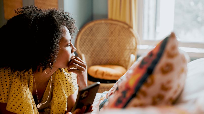 A woman thinking about home insurance with a cellphone in her hands while lying down on her bed. 