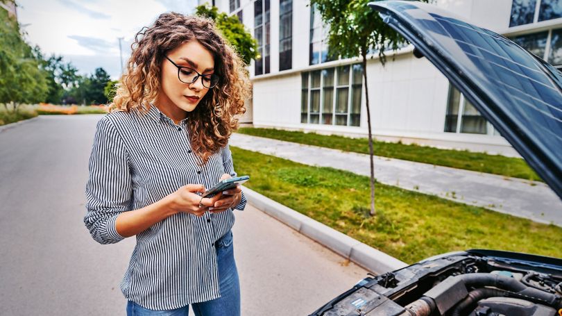 A woman using a cellphone beside a broken-down car on the side of the road. 