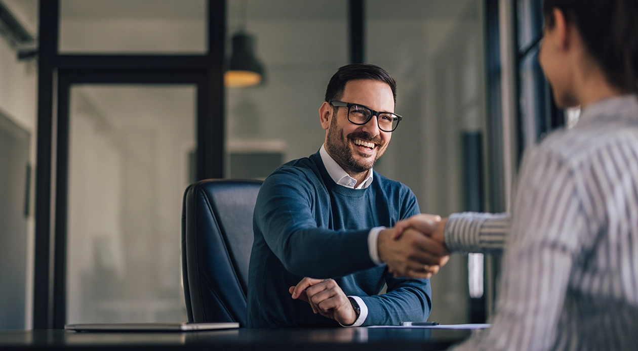 Two people shaking hands across a desk during a business meeting.