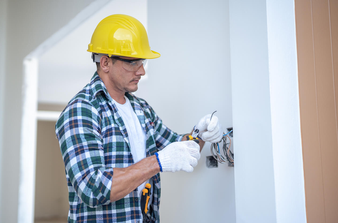 Électricien portant un casque de sécurité et des gants pendant des travaux de câblage électrique résidentiel à l’intérieur d’une maison.