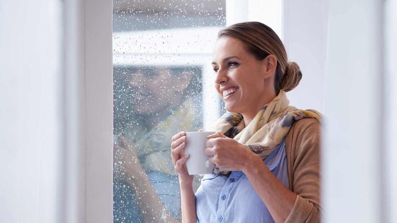 Woman holding a mug looking outside a window on a rainy day with a smile.