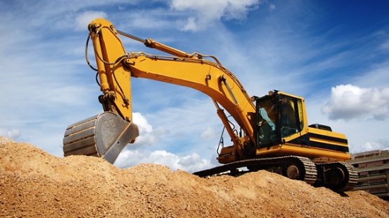 Yellow backhoe on dirt hill with building under construction in the background.