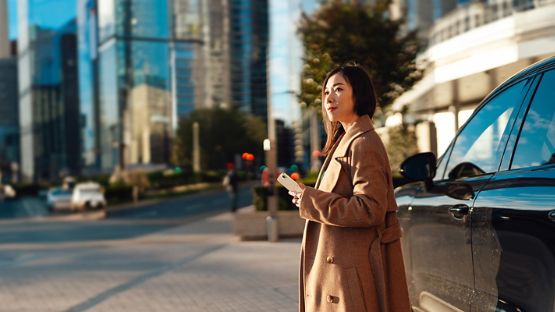 A woman locking her car with a smartphone app in front of her parked car against an urban cityscape.