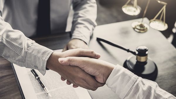 Gros plan sur un avocat serrant la main d’un client, un maillet et la balance de la* justice sont posés sur le bureau autour duquel ils se trouvent.