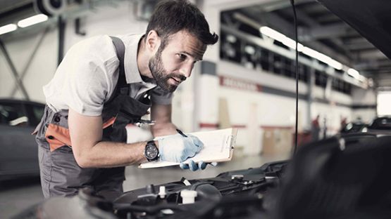 An auto repair expert with a clipboard inspecting under the hood of a vehicle.