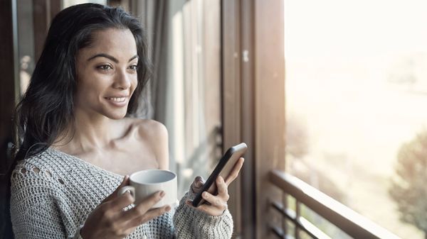 Une femme buvant un café devant une fenêtre et tenant un cellulaire.