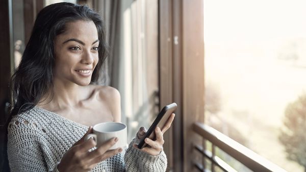 Woman standing near window drinking coffee and using her cellphone.