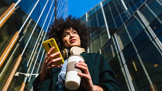 Image of woman in her 20s looking at her phone and holding a coffee
