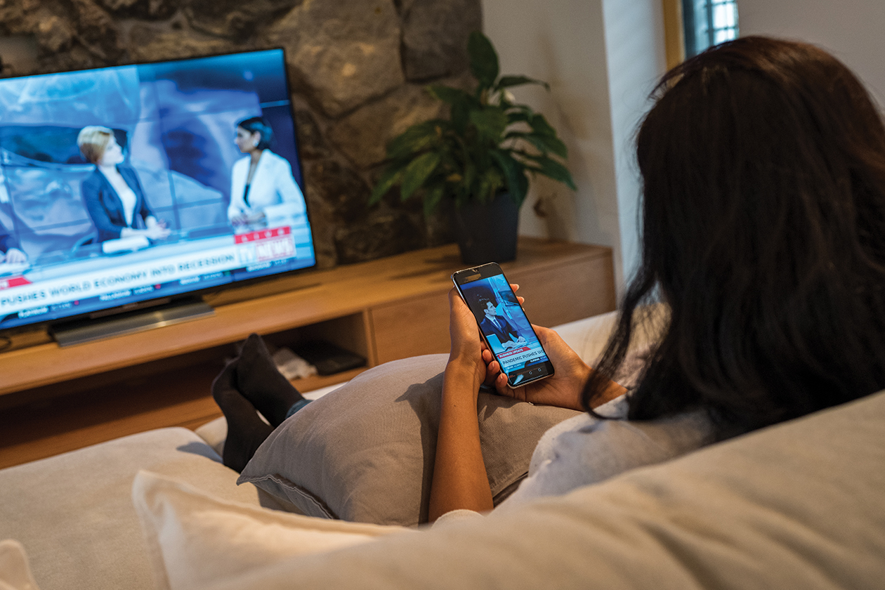 A woman sitting on a sofa watching the local news on her television while checking her emergency alerts on her phone.